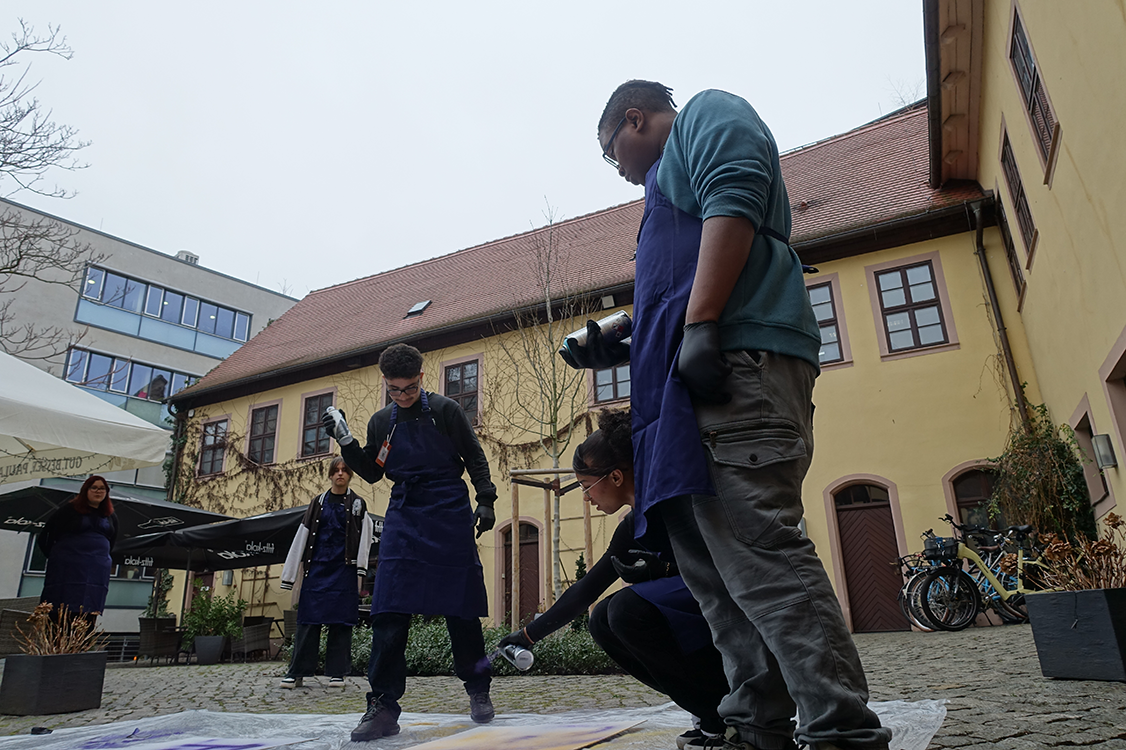 eine Gruppe aus 4 Personen steht in einem Innenhof und besprühen ein großes Papier mit Spraydosen