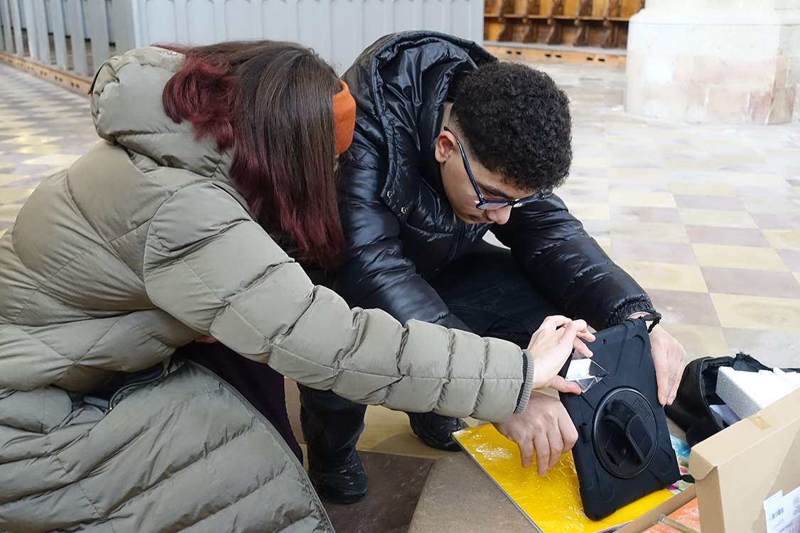 eine junge Frau und ein junger Mann hocken auf dem Boden und machen Fotos mit einem Tablet