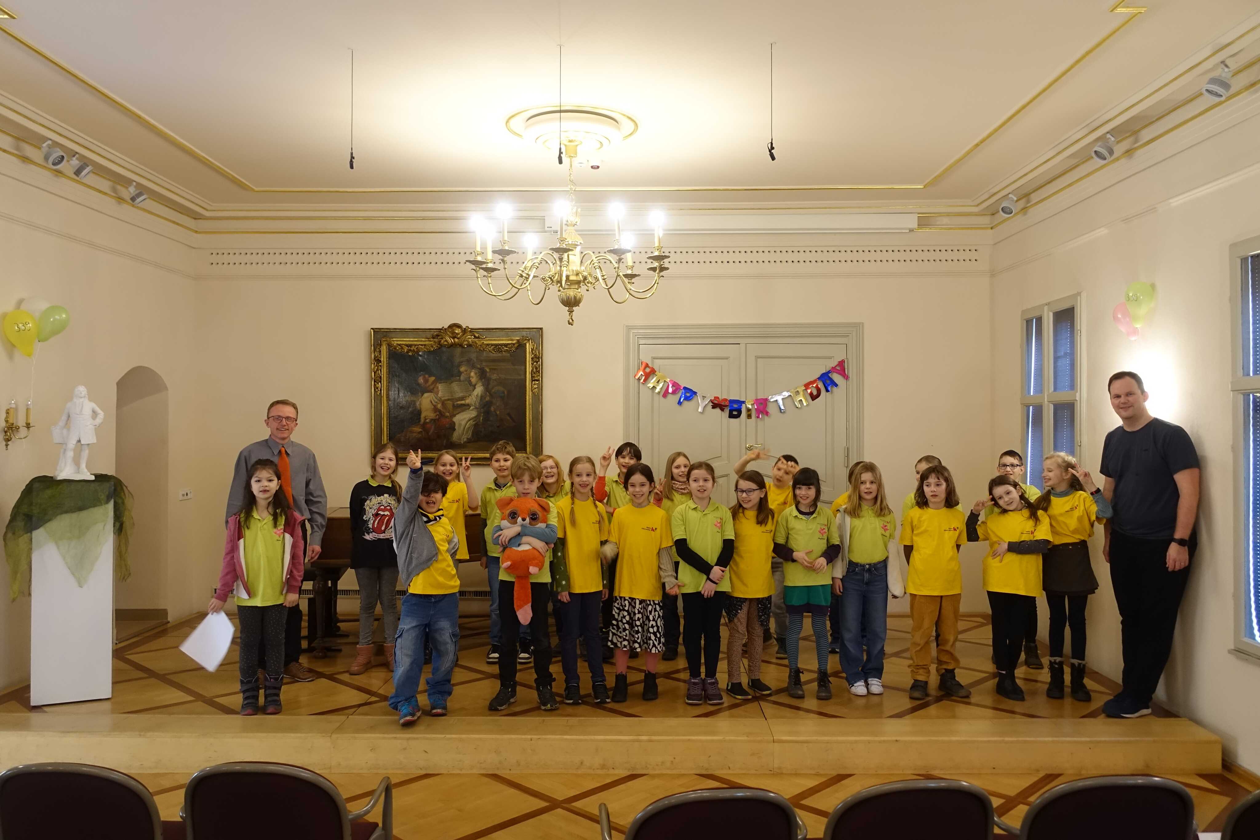 eine Gruppe von Kindern mit gelben T-Shirts sowie zwei Erwachsene stehen auf einer Bühne, im Hintergrund ist eine Geburtstagsgirlande zu sehen