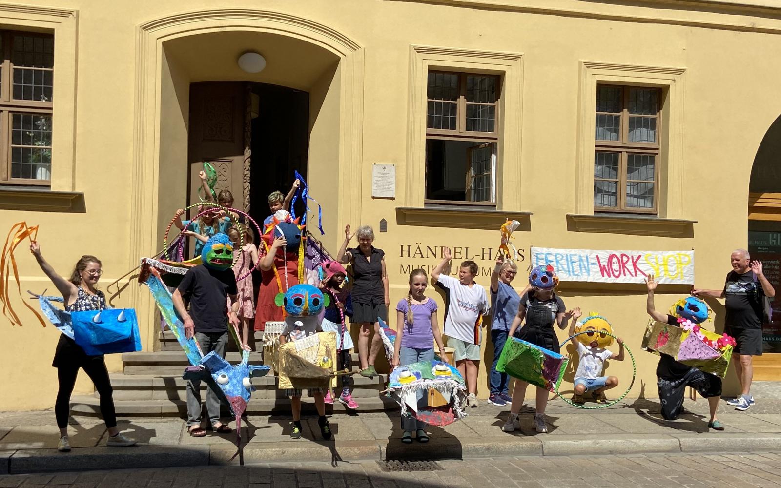 eine Gruppe Kinder stehen vor einem gelben Haus, sie tragen bunte selbstgebastelte Kostüme und Masken