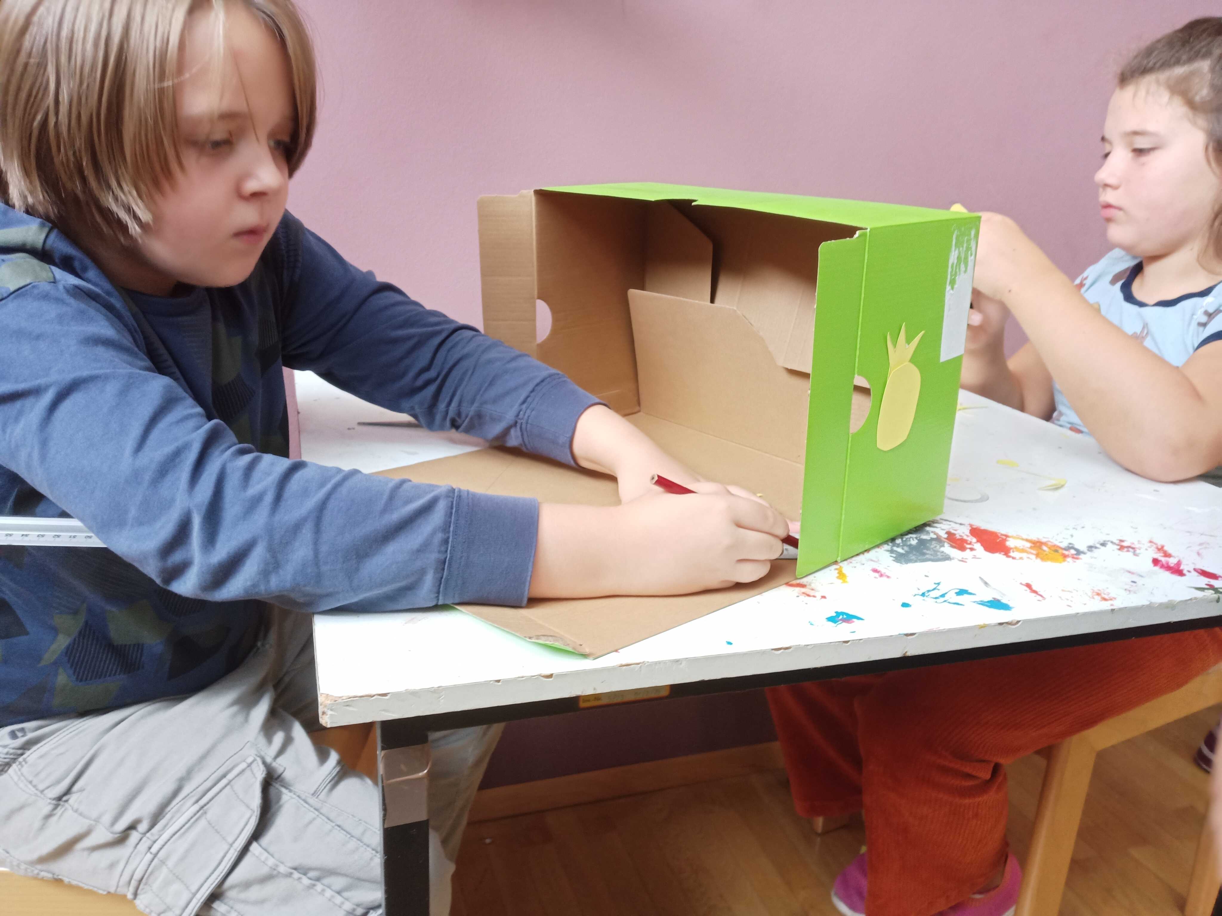 zwei Kinder sitzen sich an einem Tisch gegenüber und basteln an einem Bühnenbild im Schuhkarton