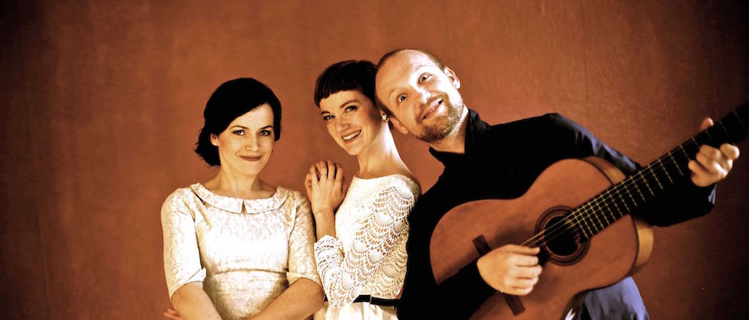 das Bild zeigt einen Mann mit einer Gitarre und zwei Sängerinnen 